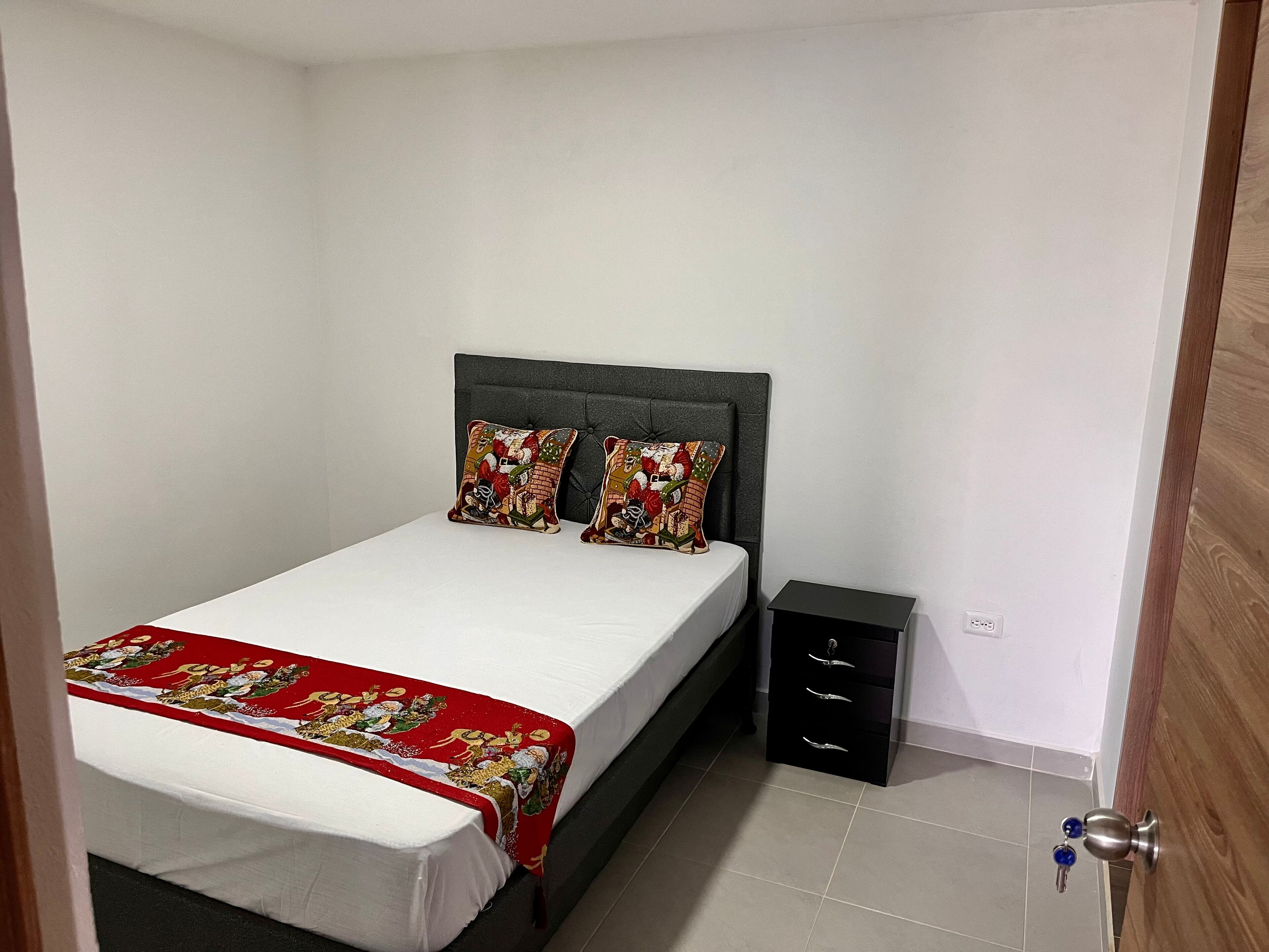 A bedroom with a bed, two decorative pillows, a bedside table, and a small drawer unit.