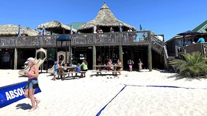 Une construction en bois située au bord de la plage, dotée d’un toit de chaume, d’un balcon et d’un sol sablonneux.