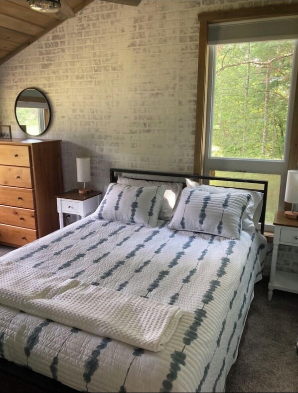 A bedroom with a bed, a dresser, a nightstand, a mirror, and a window with a view of trees.