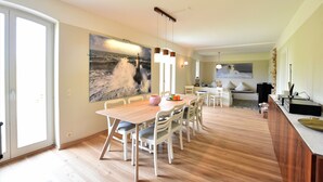 A spacious dining area with a wooden table and chairs, a large painting of a stormy sea, and a kitchen in the background. - "Haus Seegang" approx. 100 sqm, close to the beach and with a large garden (Neubukow)