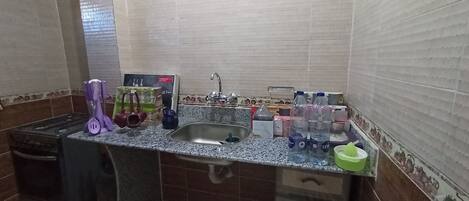 Una cocina con estufa a gas, lavamanos y diversos utensilios de cocina. Hay dos botellas de agua y un plato verde en la encimera.
