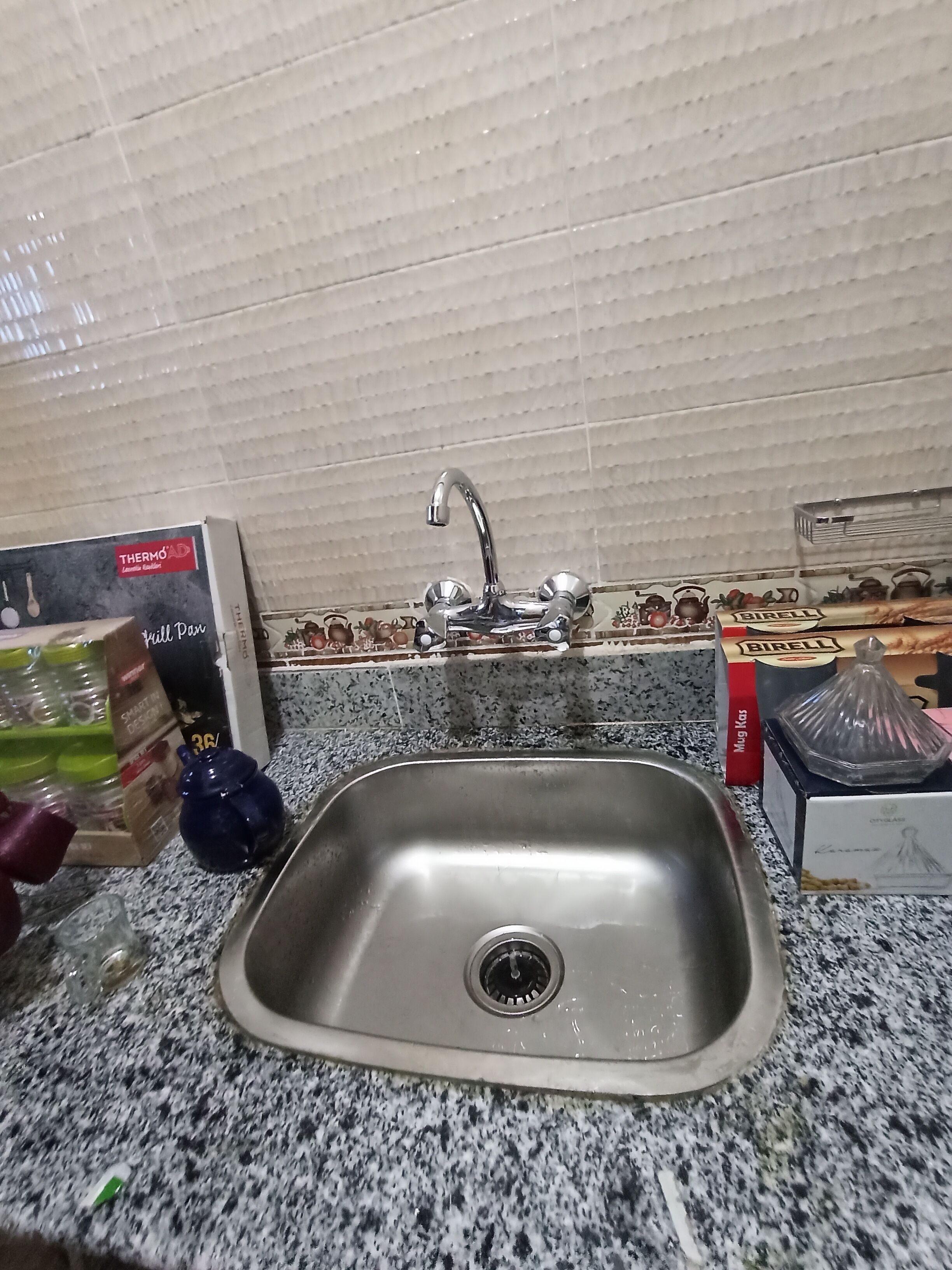 Una cocina con un lavamanos de acero inoxidable, una caja etiquetada 'Thermo' a la izquierda y una caja etiquetada 'Mug Kosh' a la derecha.