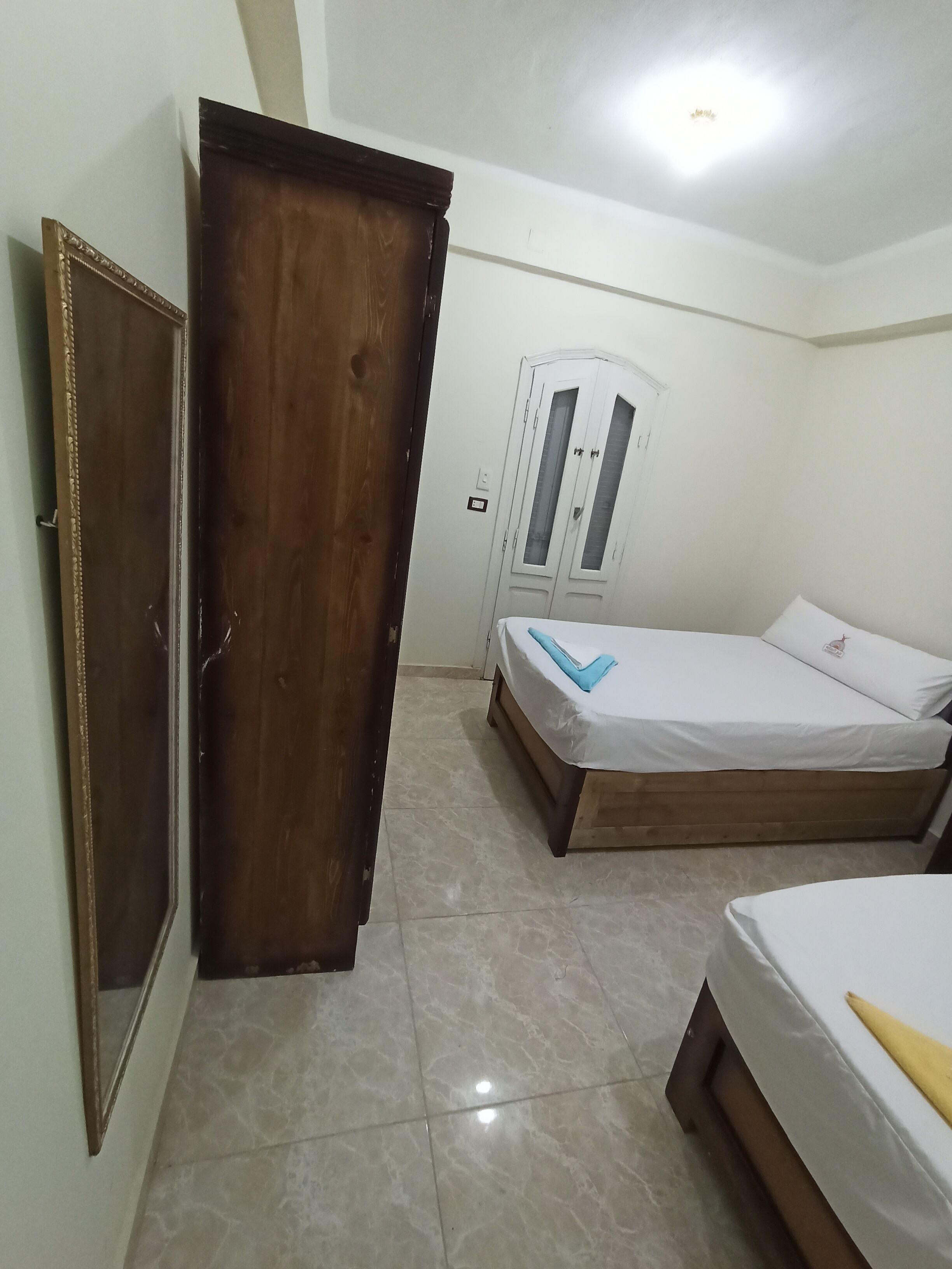 A bedroom with a wooden wardrobe, a bed with a white mattress and blue pillow, and a second bed with a white mattress and yellow pillow.