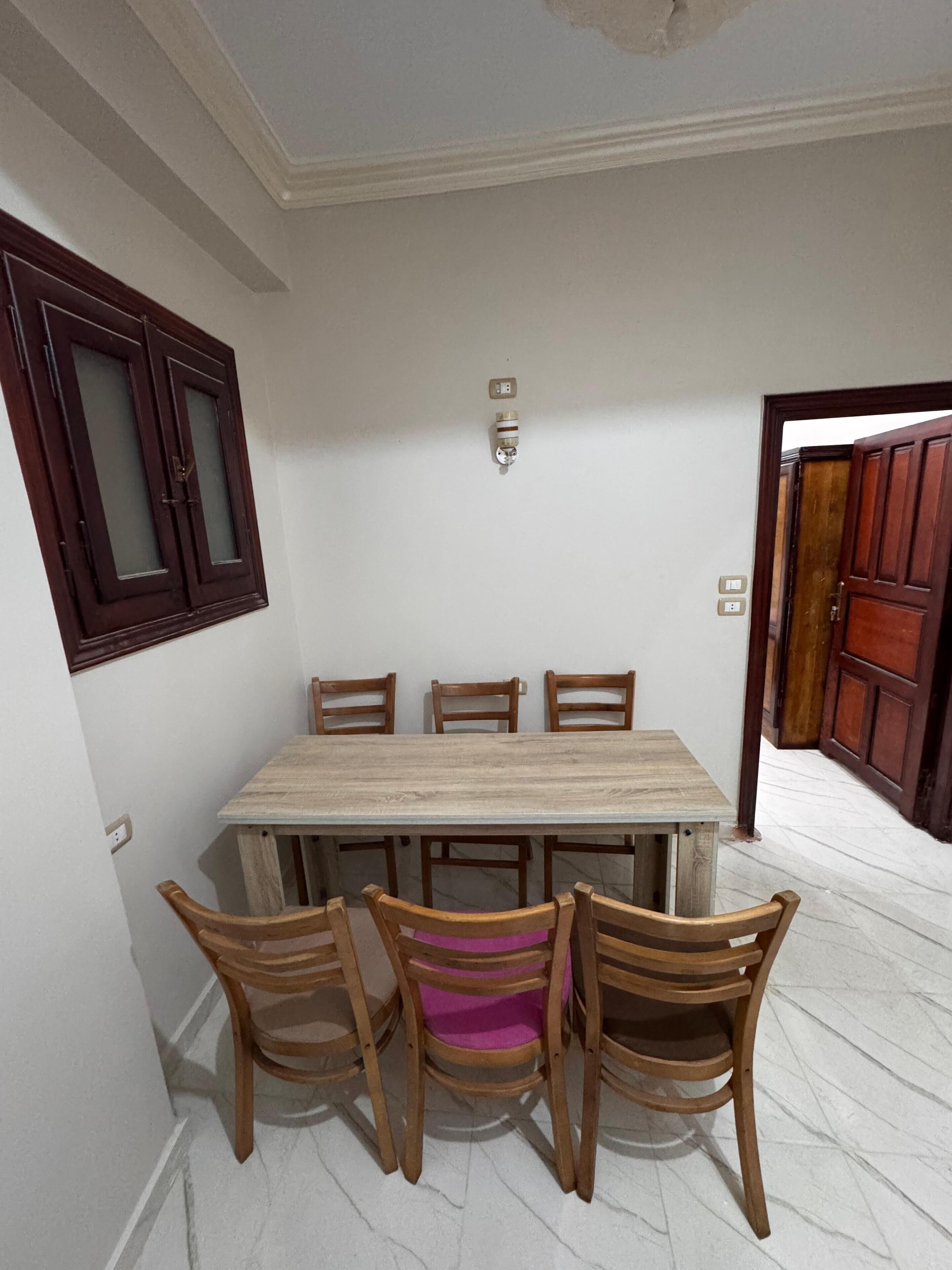 A dining area with a rectangular table and four wooden chairs. There is a wall-mounted light fixture above the table and a wooden door with glass panels to the right.