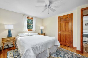 Un dormitorio con una cama, dos lámparas de noche, un ventilador de techo, un ventanal con cortinas, una puerta de madera y un tapete estampado.
