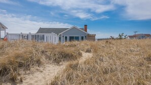 A beach house with a sandy foreground, a white fence, and a clear blue sky. - Cozy Sagamore Beach Cottage w/ Modern Comforts (Sagamore Beach)