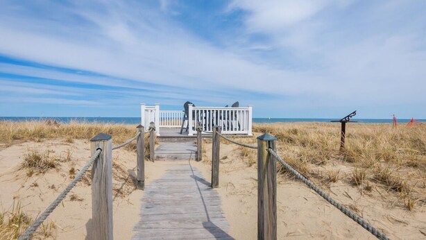 A wooden boardwalk leading to a white beachside structure with a railing, surrounded by sand and dunes. - Cozy Sagamore Beach Cottage w/ Modern Comforts (Sagamore Beach)