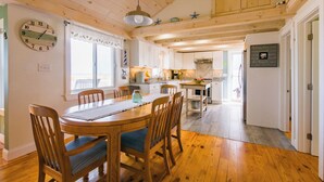 A wooden dining table with chairs, a clock, and a window with curtains. - Cozy Sagamore Beach Cottage w/ Modern Comforts (Sagamore Beach)
