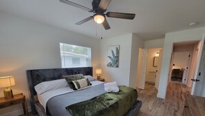 Un dormitorio con cama, ventilador de techo, ventana, mesita de noche y puerta que da a otra habitación.