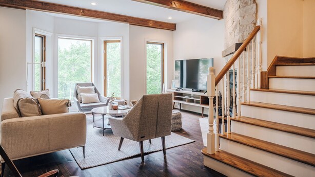 A spacious living room with a large TV, wooden staircase, and a stone wall. - 5BD Scani Chic! Mountain View! 6 mins to Skiing! (Mont-Tremblant)