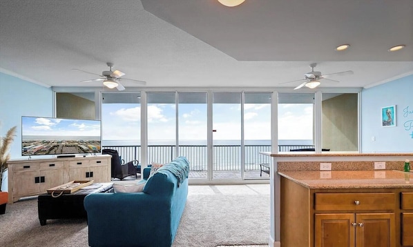 A spacious living area with a large window overlooking the ocean, a blue sofa, a TV, and a kitchen area with wooden cabinets.
