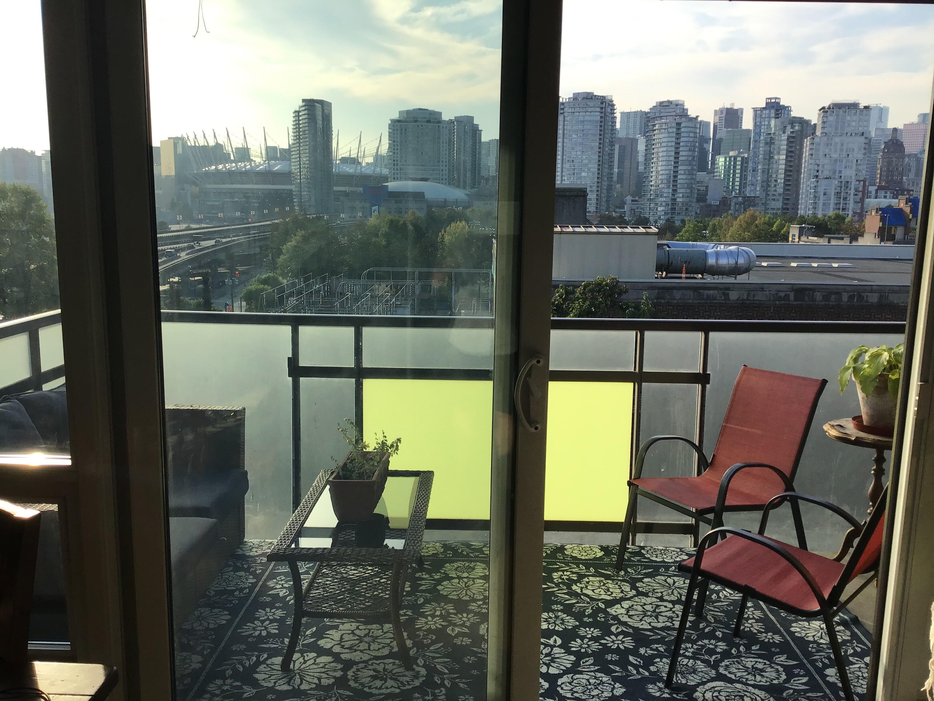 A balcony with a view of a city skyline, a table with a potted plant, and two red chairs.