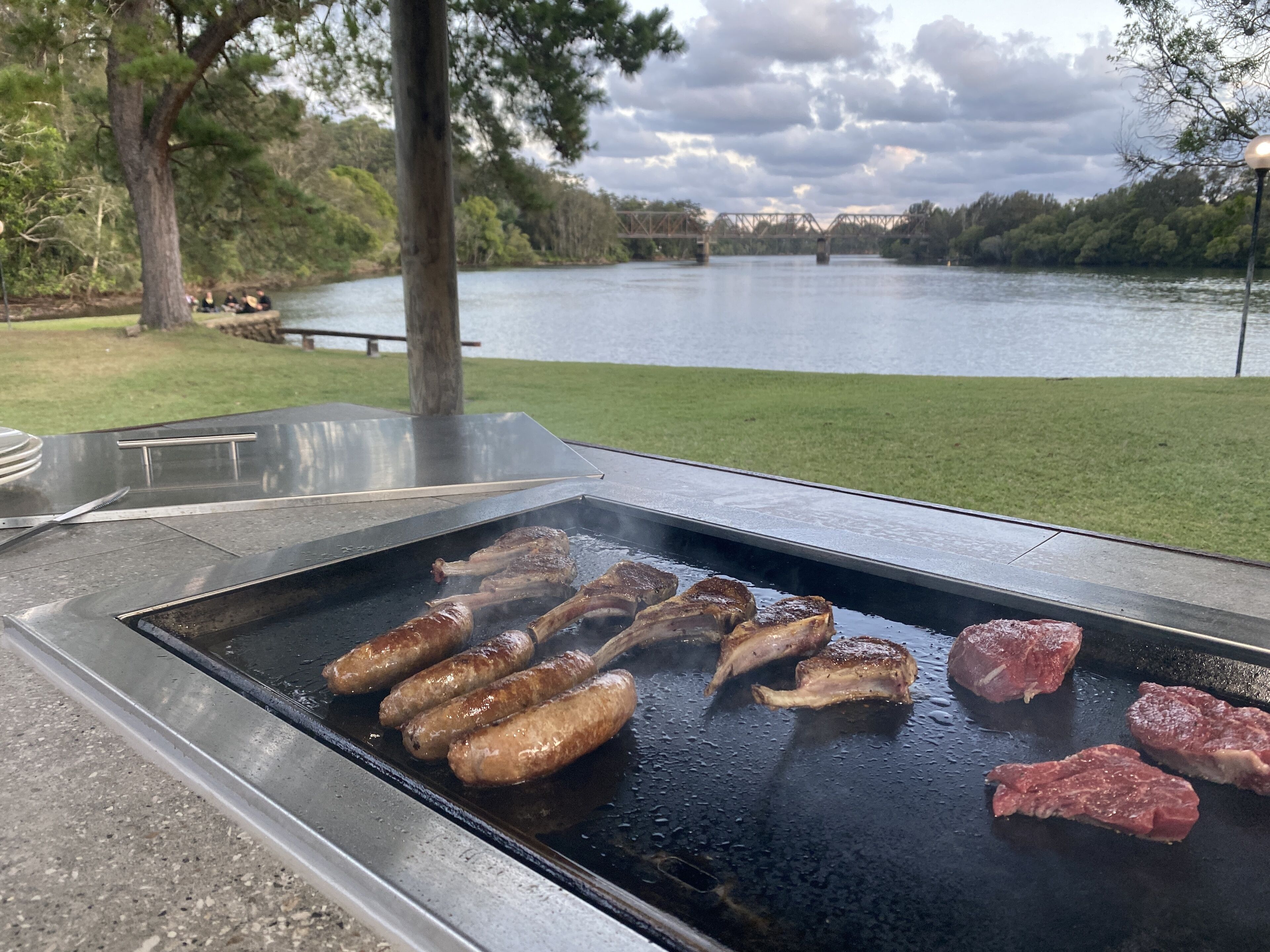 Ein Grill im Freien mit Bratwürsten und Steaks, umgeben von einer Rasenfläche und einem See mit einer Brücke im Hintergrund.