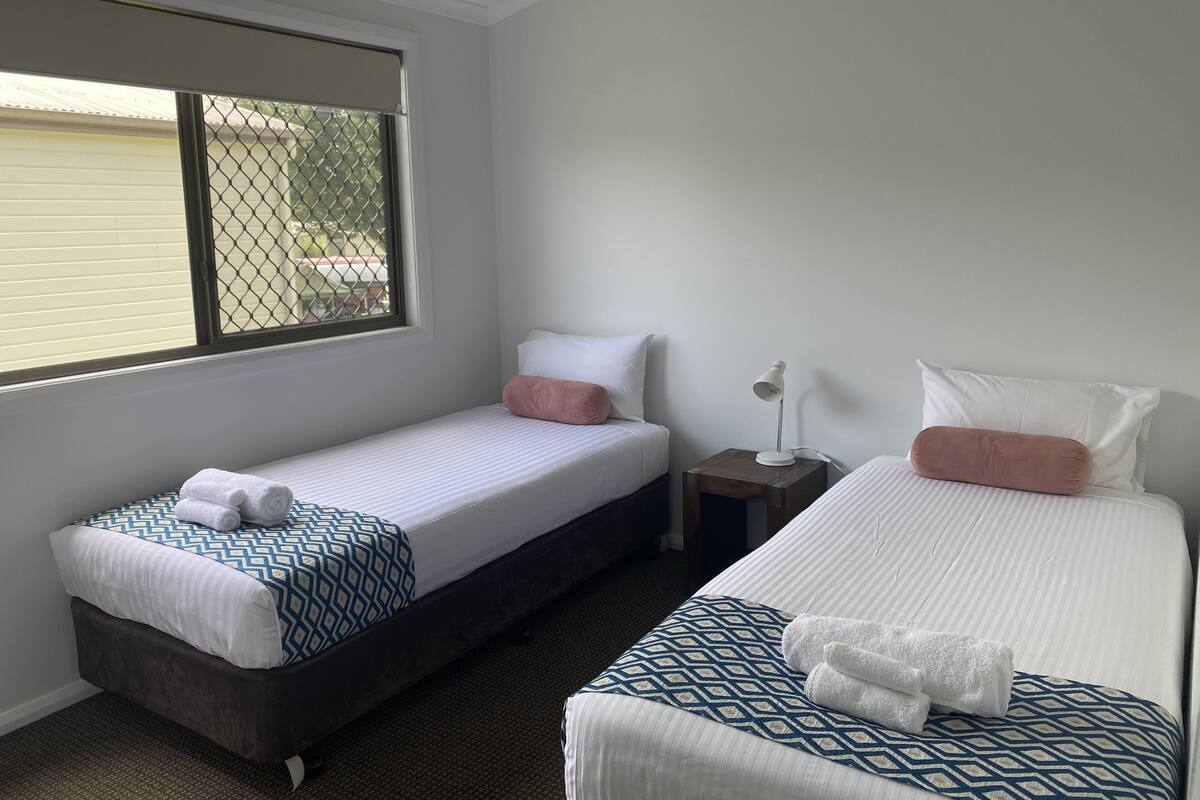 Ein Hotelzimmer mit zwei Einzelbetten, einem kleinen Nachttisch mit Lampe und einem Fenster mit Metallgitter.