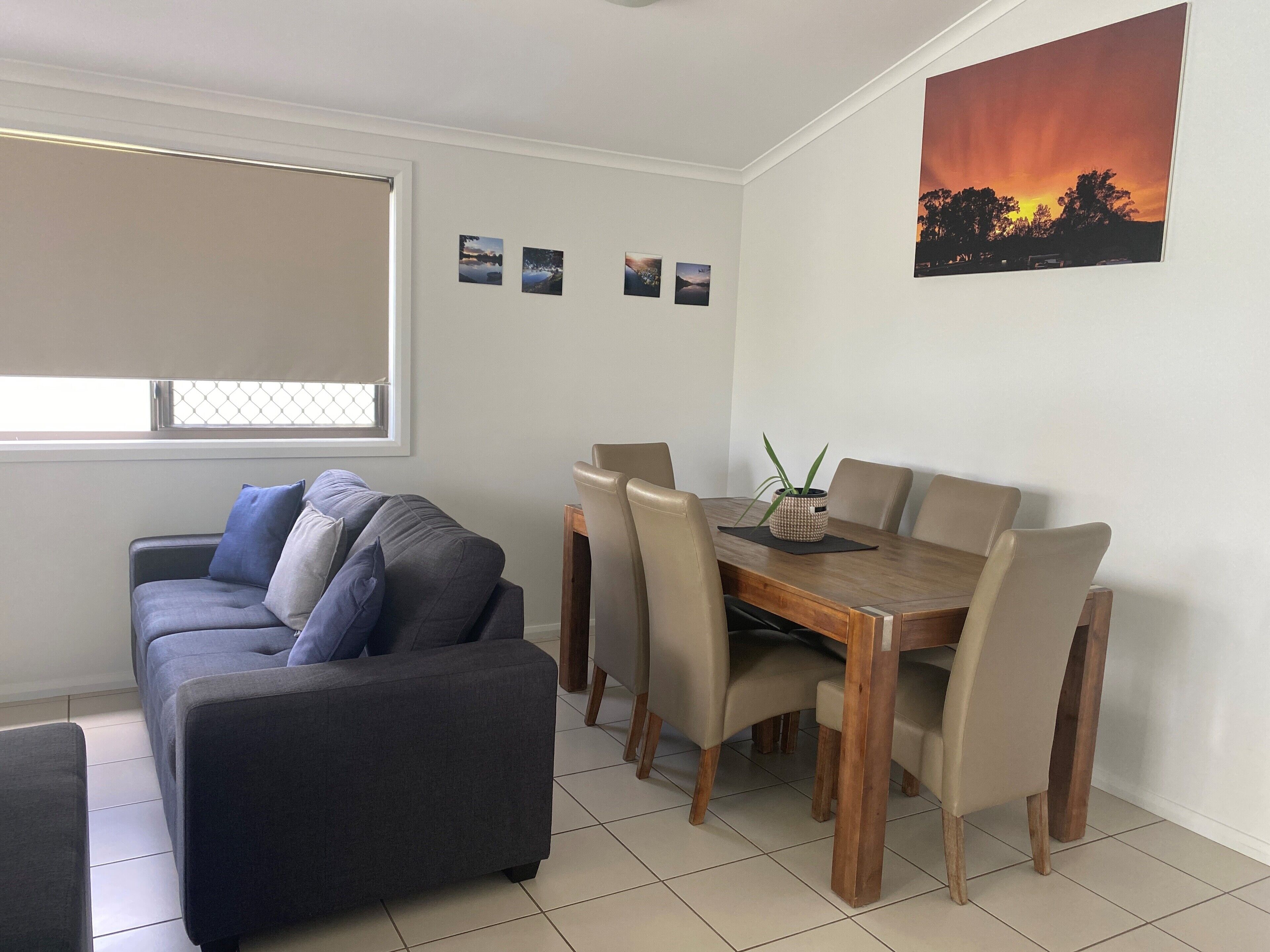 Een woonkamer met een bank, een eettafel met stoelen, een raam met zonwering en een schilderij van een zonsondergang aan de muur.