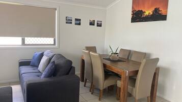 Een woonkamer met een bank, een eettafel met stoelen, een raam met zonwering en een schilderij van een zonsondergang aan de muur.
