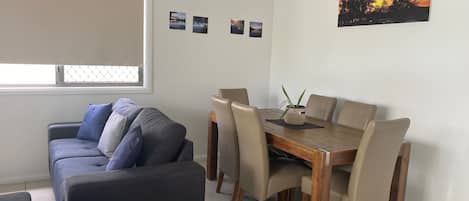 Een woonkamer met een bank, een eettafel met stoelen, een raam met zonwering en een schilderij van een zonsondergang aan de muur.