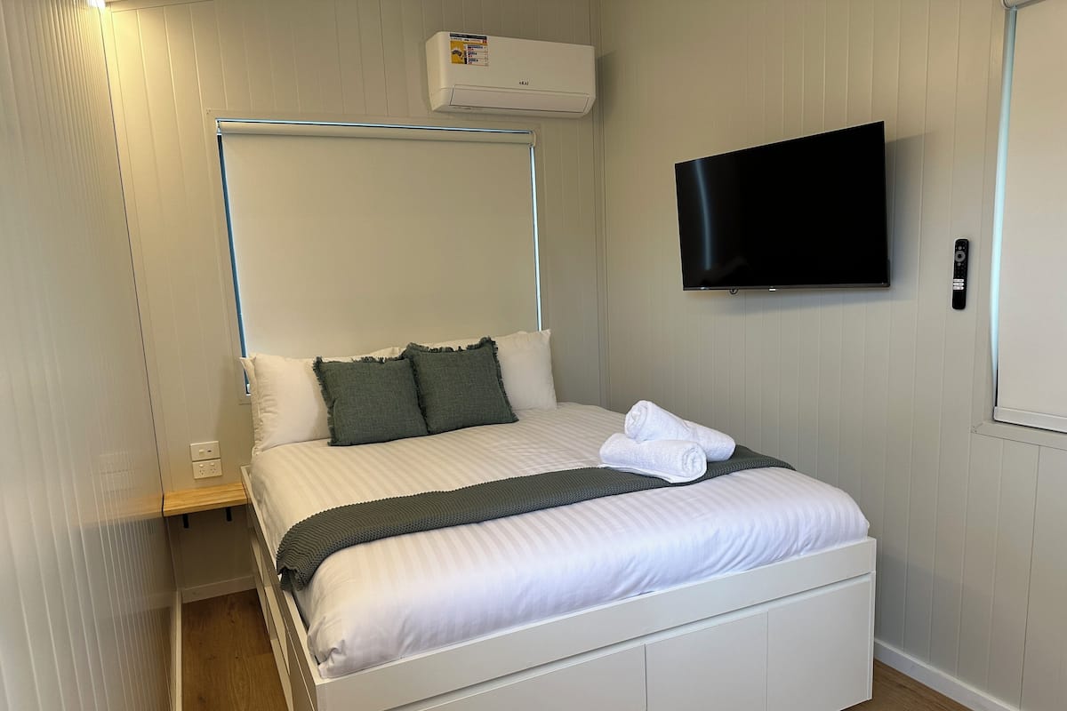 Ein ordentlich gemachtes Bett mit weißer Bettwäsche und einem dunkelgrünen Überwurf, ein an der Wand montierter Flachbildfernseher und ein kleiner Nachttisch mit Steckdose.