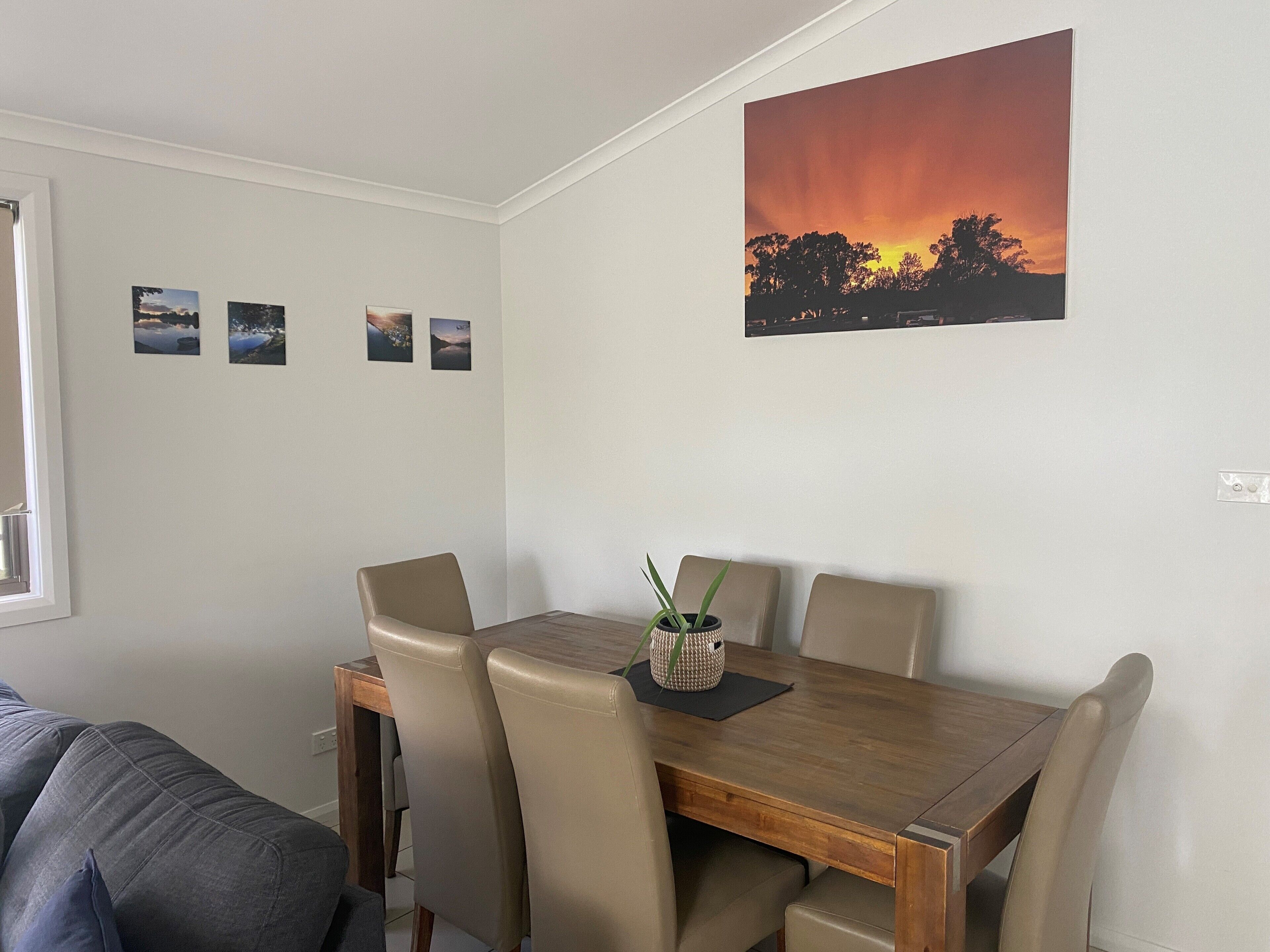A dining area with a wooden table and four chairs, a potted plant, and a sunset painting on the wall.