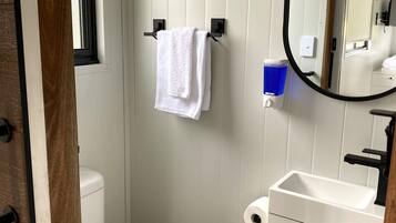 A compact bathroom with a white toilet, a towel hanging on a black towel rack, a mirror, and a sink with a soap dispenser.