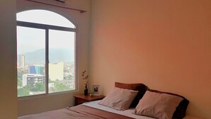 Una camera da letto con un letto, una ventola a soffitto, un condizionatore, un comodino, una finestra con vista sulla città e un vaso con fiori.