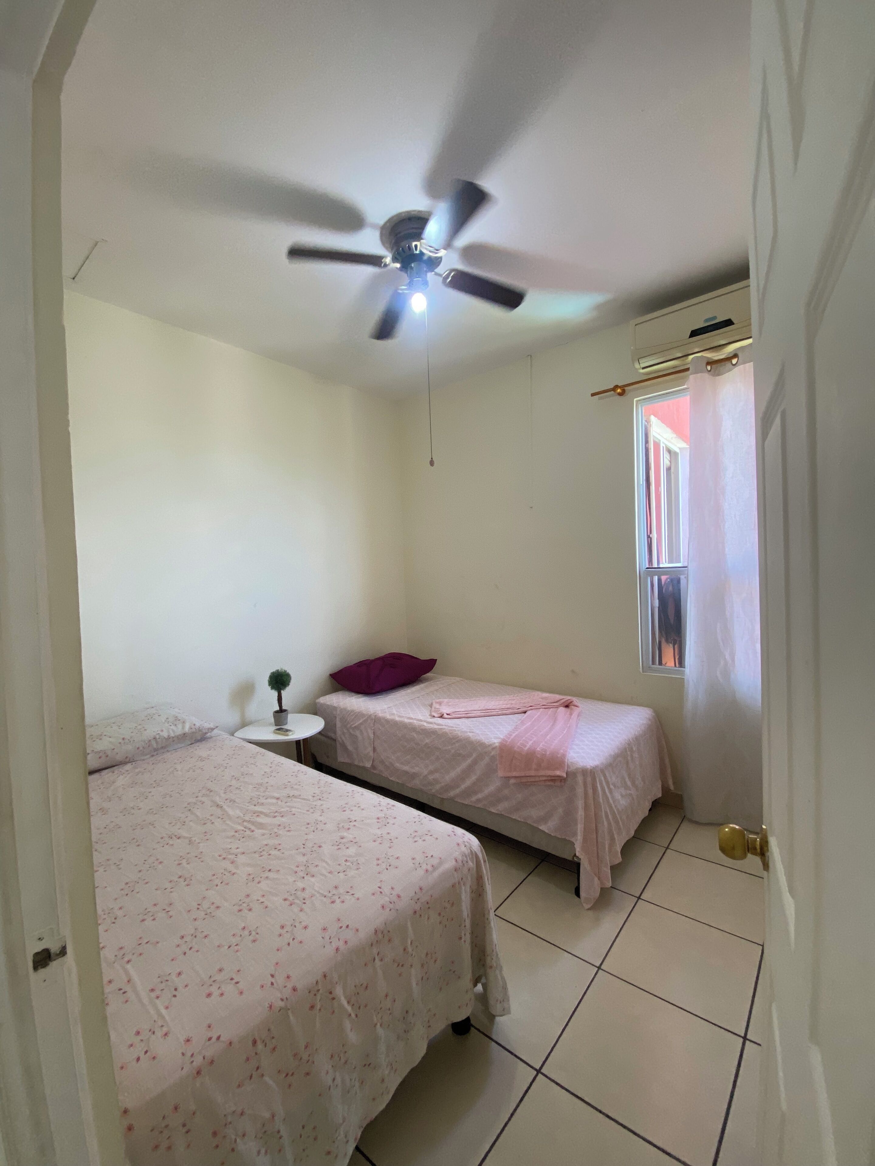 Ein kleines, sauberes Zimmer mit zwei Betten, einem Deckenventilator, einem Fenster mit Vorhängen und einem Tisch mit einer Pflanze.