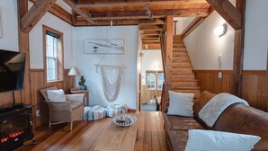 Un salón acogedor con vigas de madera, chimenea, sofá, sillón, un macramé colgante y una escalera que lleva a otra habitación.