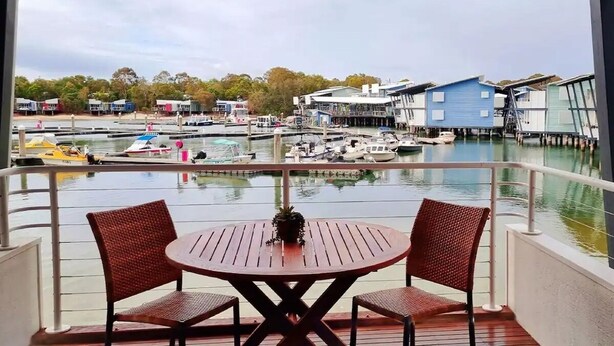 Ein Balkon mit zwei Korbstühlen und einem kleinen Tisch, der auf einen Yachthafen mit Booten und Gebäuden am Wasser blickt.