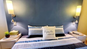 Ein ordentlich bezogenes Bett mit gestreifter Bettwäsche und einem Kissen mit der Aufschrift „Genieße den Moment“. Zwei Nachttischlampen sind an, und auf dem Nachttisch steht eine kleine Pflanze.