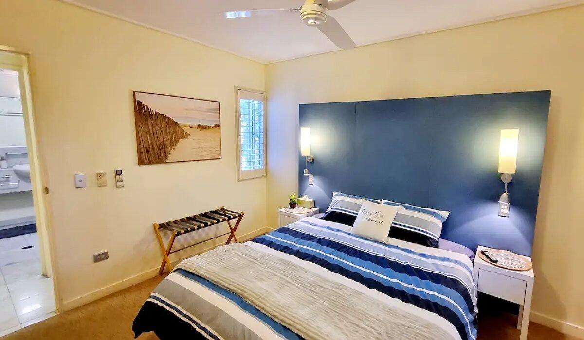 Ein Schlafzimmer mit Bett, Kopfstütze, Nachttischlampen, einem Nachttisch, einem Wandbild und einem Fenster mit Jalousien.