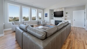 Ein modernes Wohnzimmer mit einer großen Ledersitzgruppe, einem Flachbildfernseher an einer Wand mit Ziegeloptik und großen Fenstern mit Blick ins Freie.