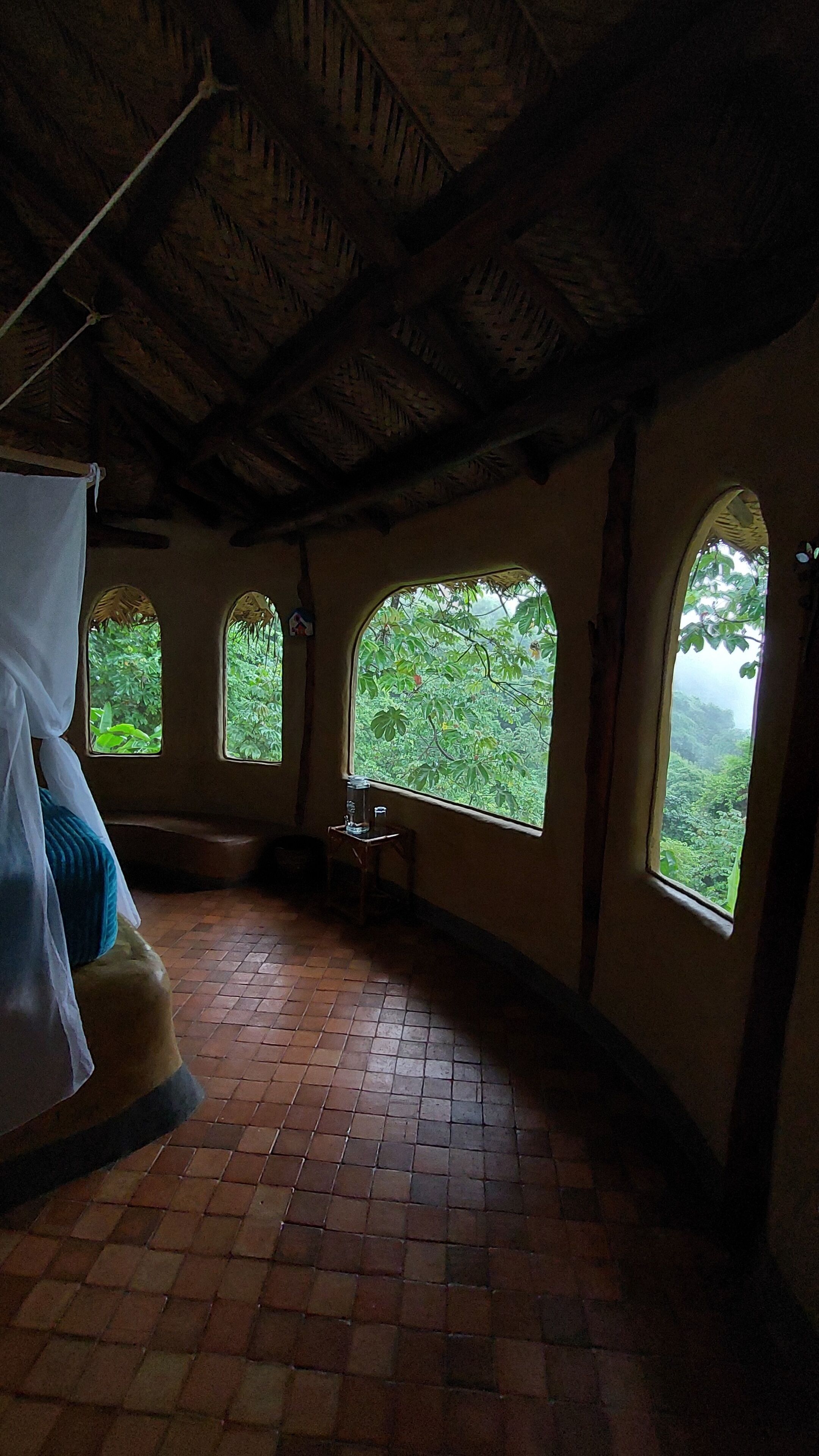 A room with a tiled floor, a bed with a canopy, and three arched windows overlooking a forested area.
