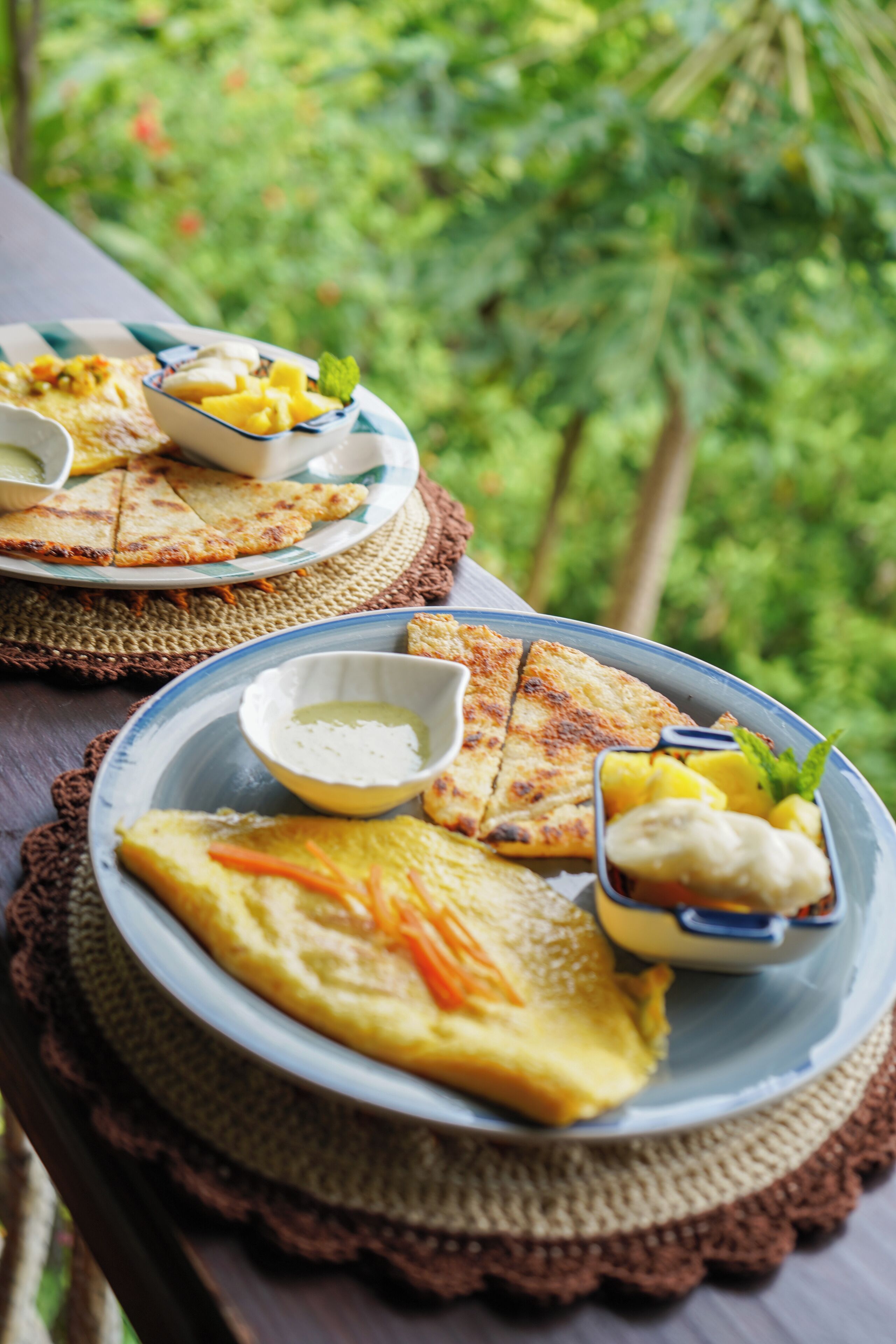 A table with plates of food, including grilled items and a bowl of sauce, set on a woven mat outdoors.