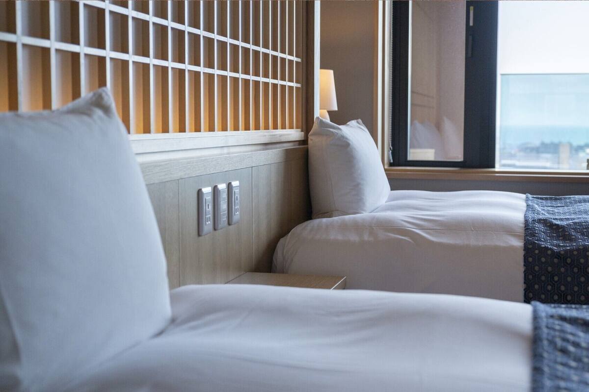 Ein Hotelzimmer mit zwei Betten, einem Fenster mit Blick auf die Stadt und einer Wand mit einer Leuchte.