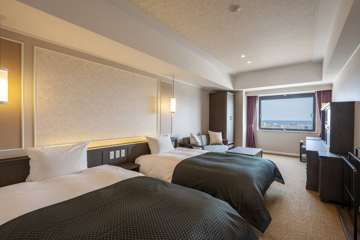 Ein Hotelzimmer mit zwei Betten, einem Schreibtisch, einem Sessel, einem Fernseher und einem großen Fenster mit Blick auf die Stadt.