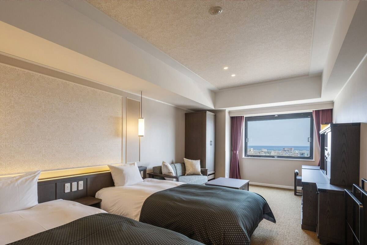 Ein Hotelzimmer mit zwei Betten, einem Schreibtisch, einem Stuhl, einem großen Fenster mit Blick auf die Stadt und einer Wand mit einer dekorativen Verkleidung.