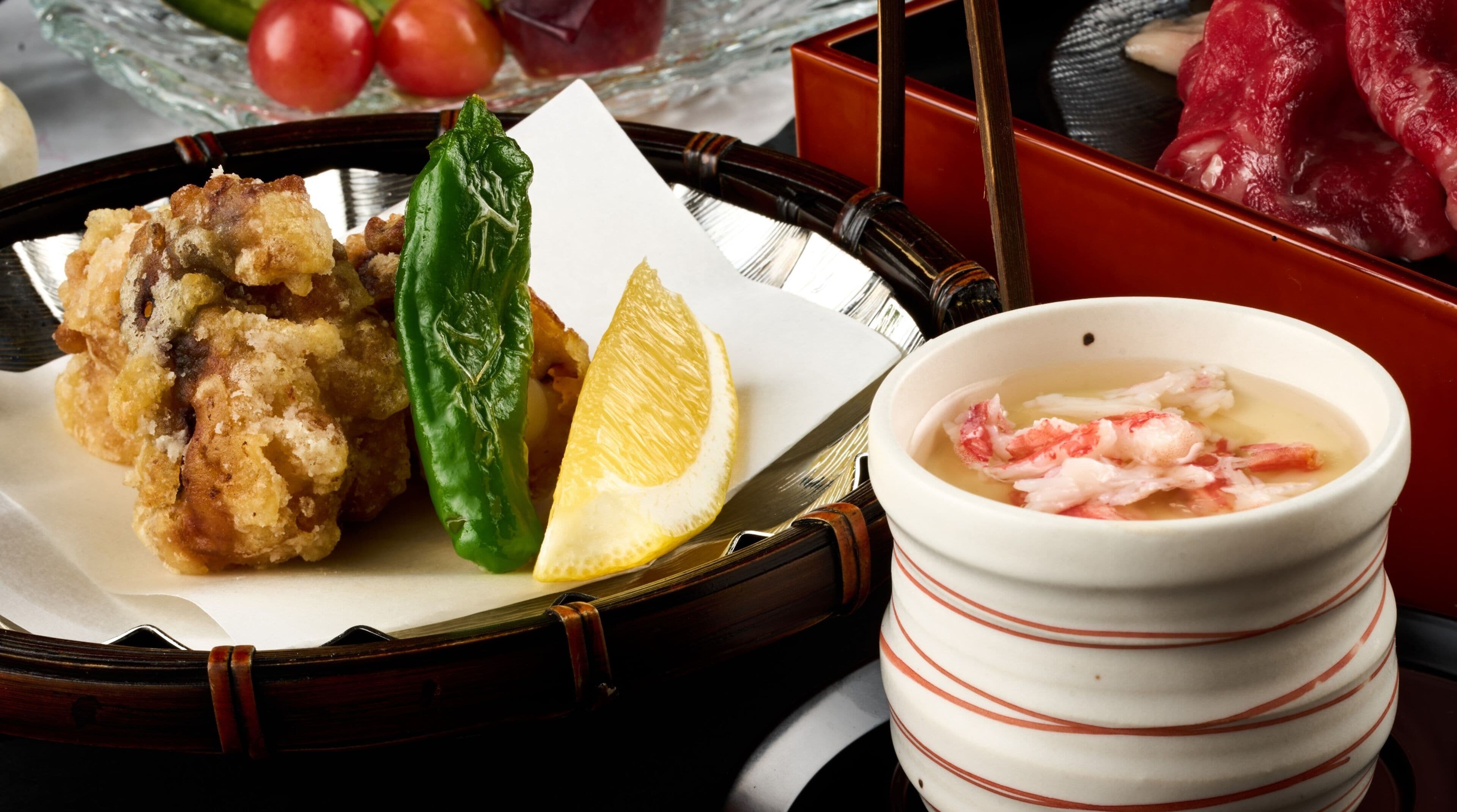 A traditional Japanese meal with tempura, lemon, and green pepper on a white plate, and a bowl of soup with seafood on the side.