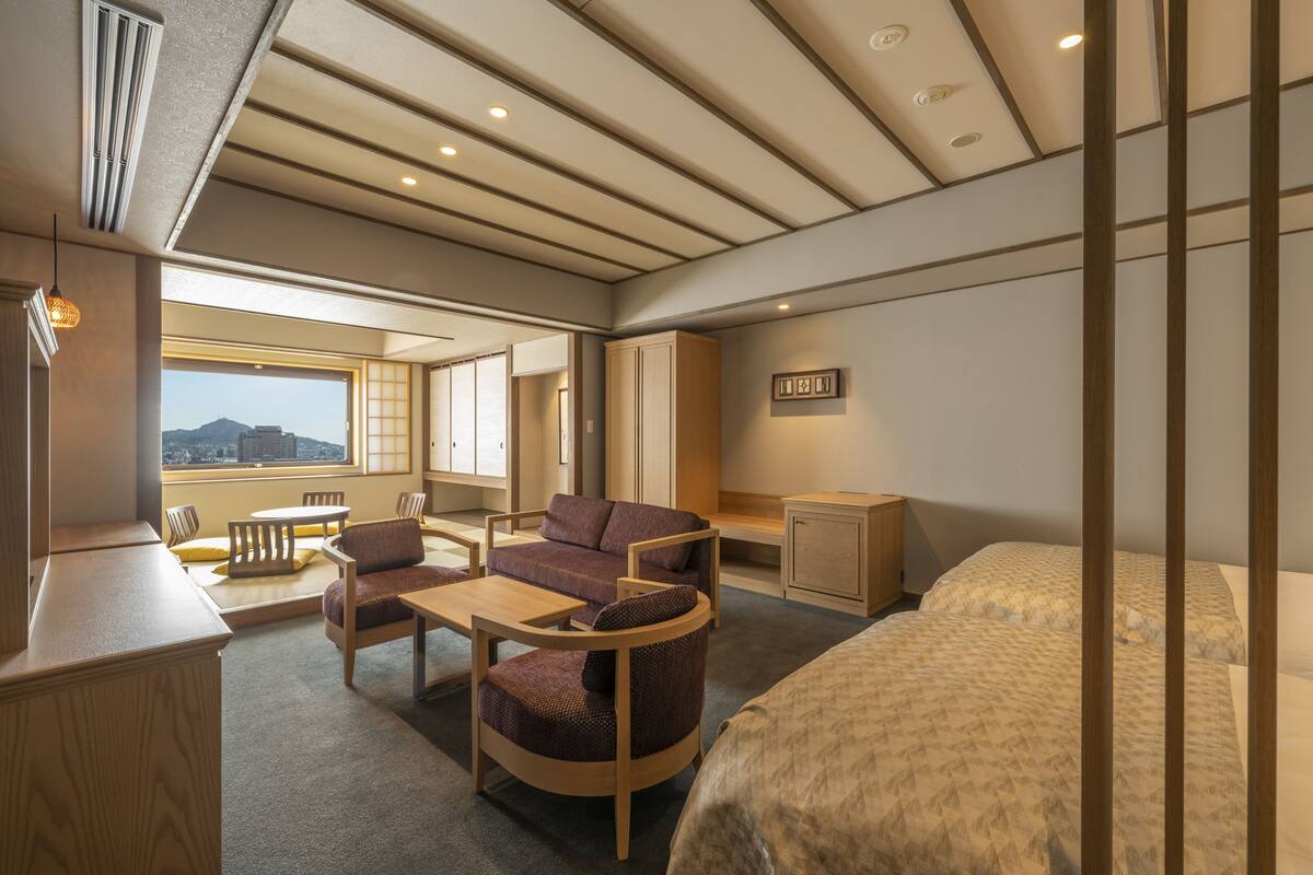 Ein modernes Hotelzimmer mit einem großen Bett, einer Sitzecke mit Sofa und Sesseln, einem kleinen Tisch und Blick auf die Stadt durch große Fenster.
