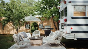 A backyard with a white RV, a patio with a woven coffee table, and a grill. - Romantic lakefront skoolie for 4: queen, twins, hot tub and private bathhouse (Granbury)