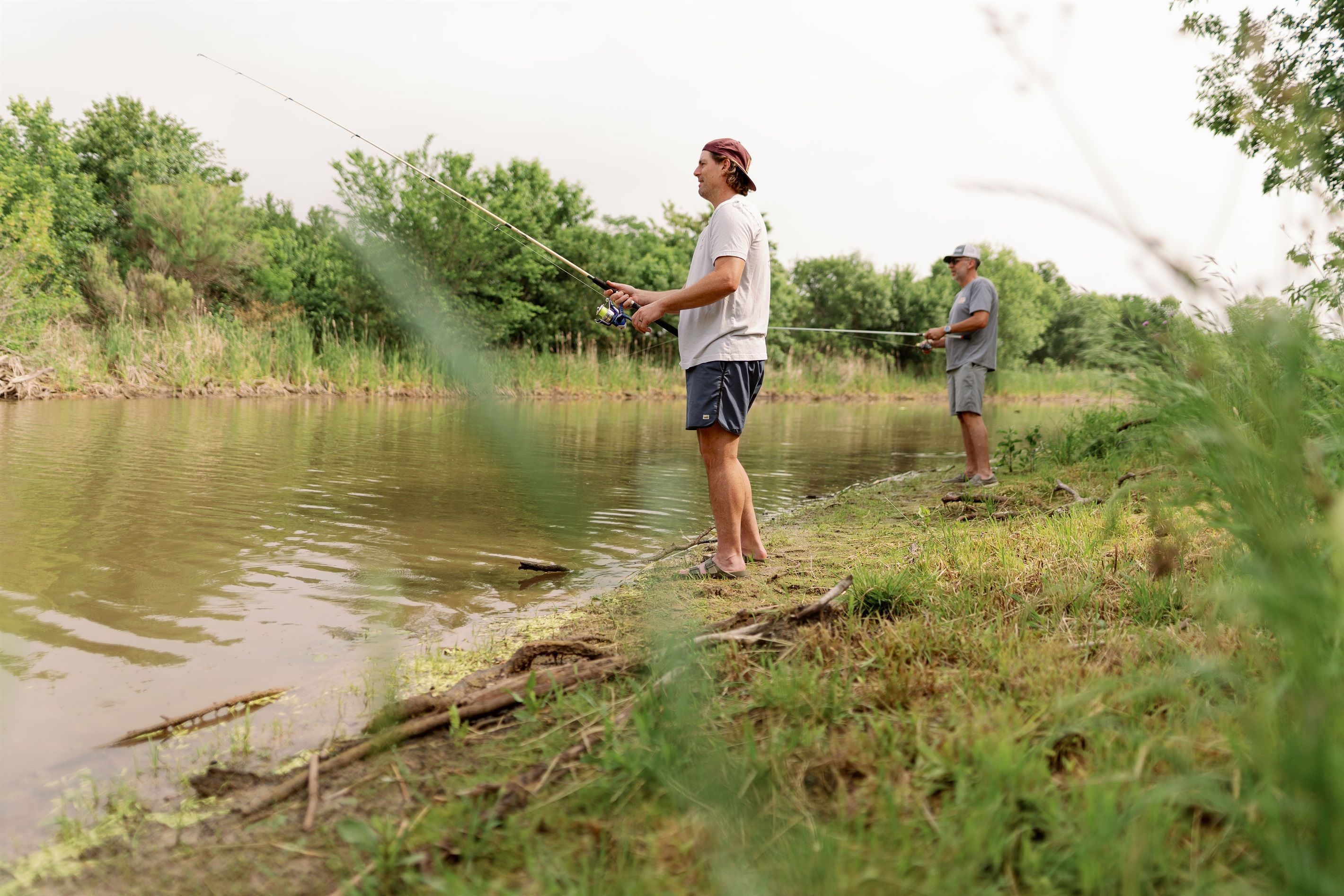 Two individuals fishing by a calm river with greenery in the background.