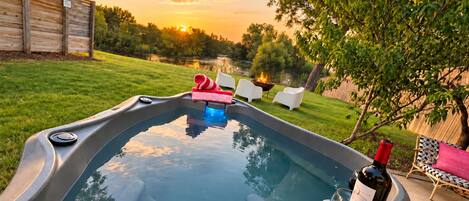 Un bain à remous agrémenté d’un foyer, entouré d’une pelouse et d’arbres, avec un coucher de soleil en arrière-plan.