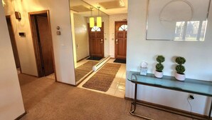 A hallway with a glass console table, two potted plants, and a mirror reflecting a door and a light fixture.