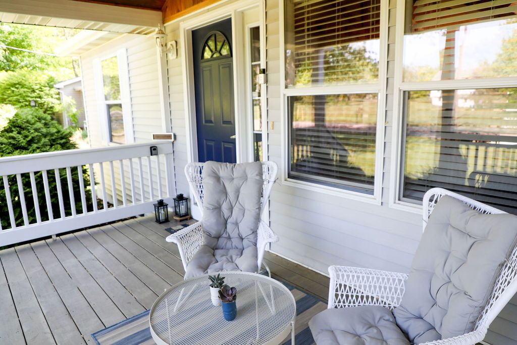 Une véranda avec des fauteuils en osier blancs et une table ronde, une porte bleue et une balustrade blanche.