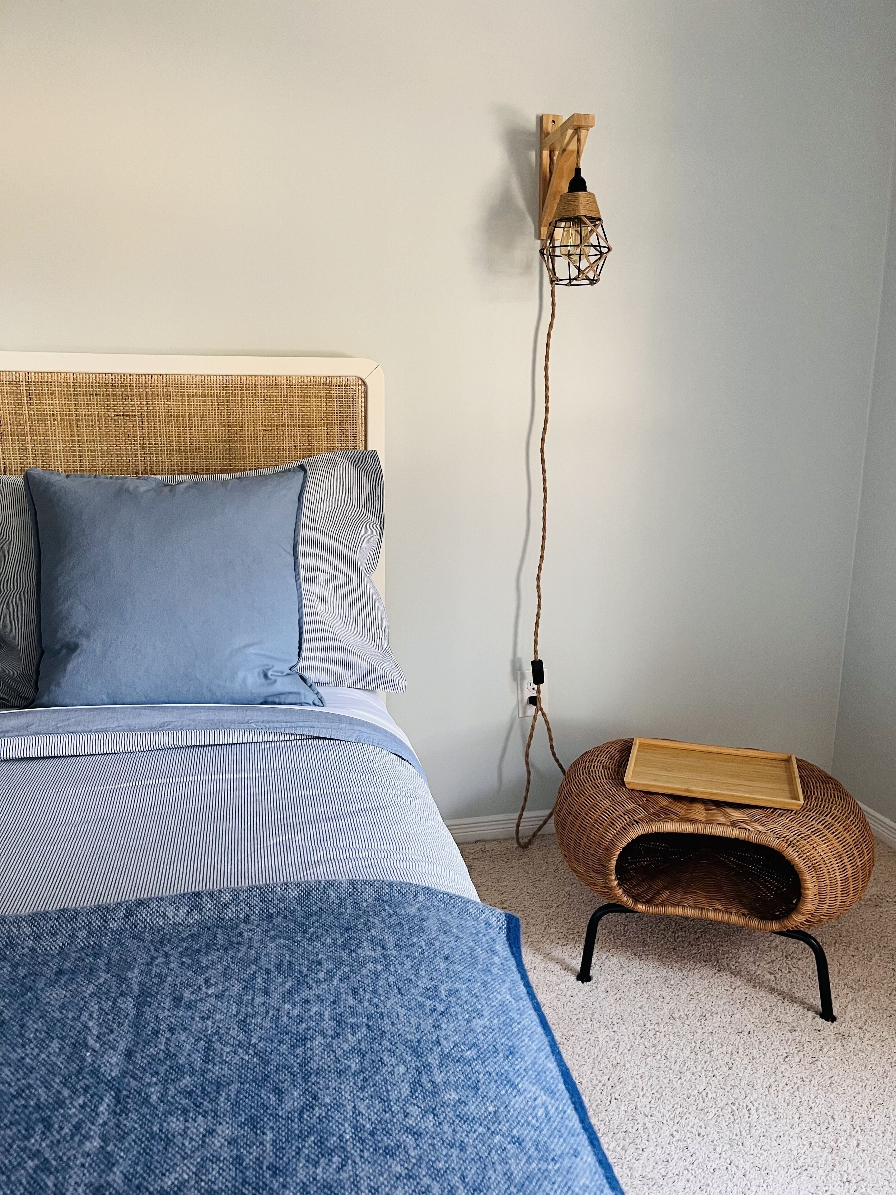 Una cama con cabecera de fibra entrelazada, almohada azul y manta a rayas. Se observa un aplique de pared y una pequeña cesta de mimbre en el suelo.