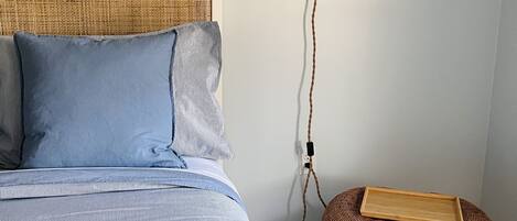 Una cama con cabecera de fibra entrelazada, almohada azul y manta a rayas. Se observa un aplique de pared y una pequeña cesta de mimbre en el suelo.