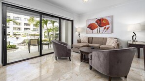 A modern hotel room with a large sliding glass door leading to a pool area, a grey sofa, a coffee table, and a painting of red flowers on the wall. - Beautiful One Bedroom Ground Level @ PalmAruba (Noord)