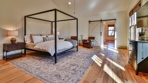 A spacious bedroom with a four-poster bed, a wooden floor, a large rug, and a view of the hallway.