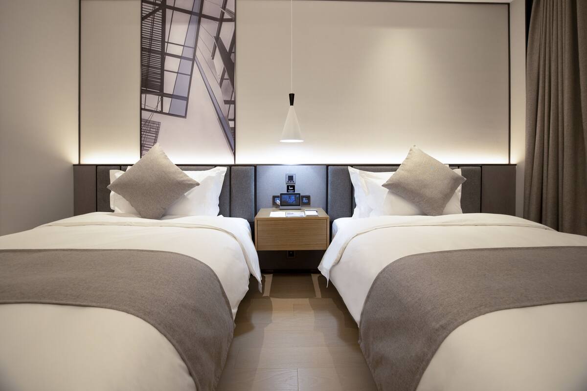 A modern hotel room with two beds, a nightstand, a digital clock, and a large window with a view of a building.