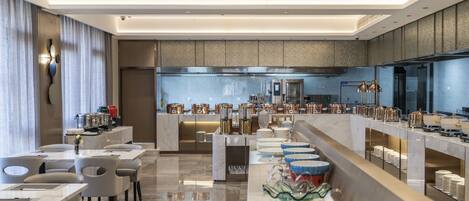 A modern hotel lobby with a long reception desk, seating area, and a kitchen in the background.