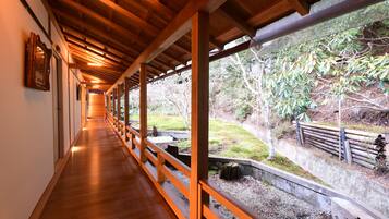 A covered wooden walkway with a railing, leading to a garden area with trees and a stone wall.